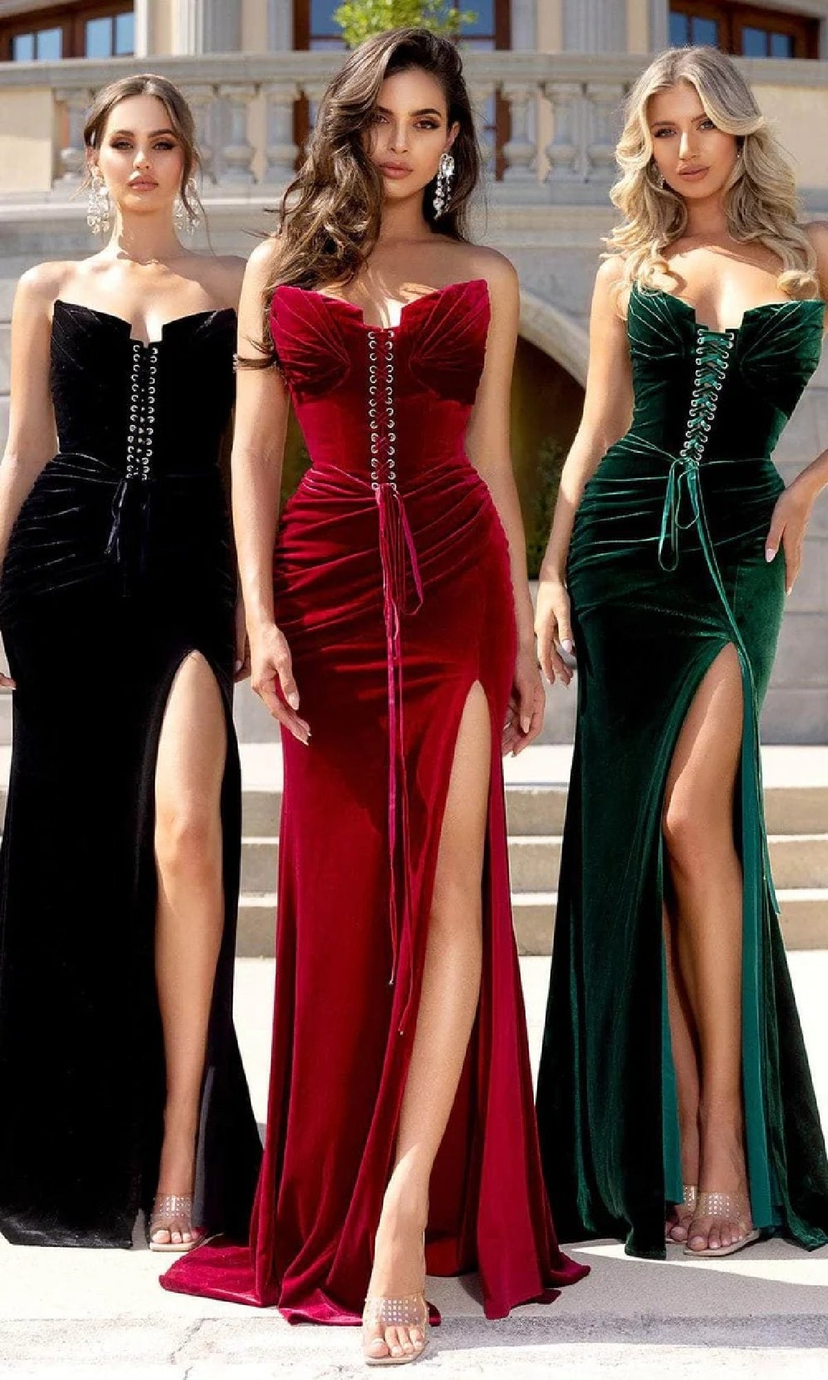 Three women wearing the Portia and Scarlett PS25640E long prom dress in black, featuring velvet fabric, thigh-high slits, and lace-up corset fronts, stand together on outdoor steps.